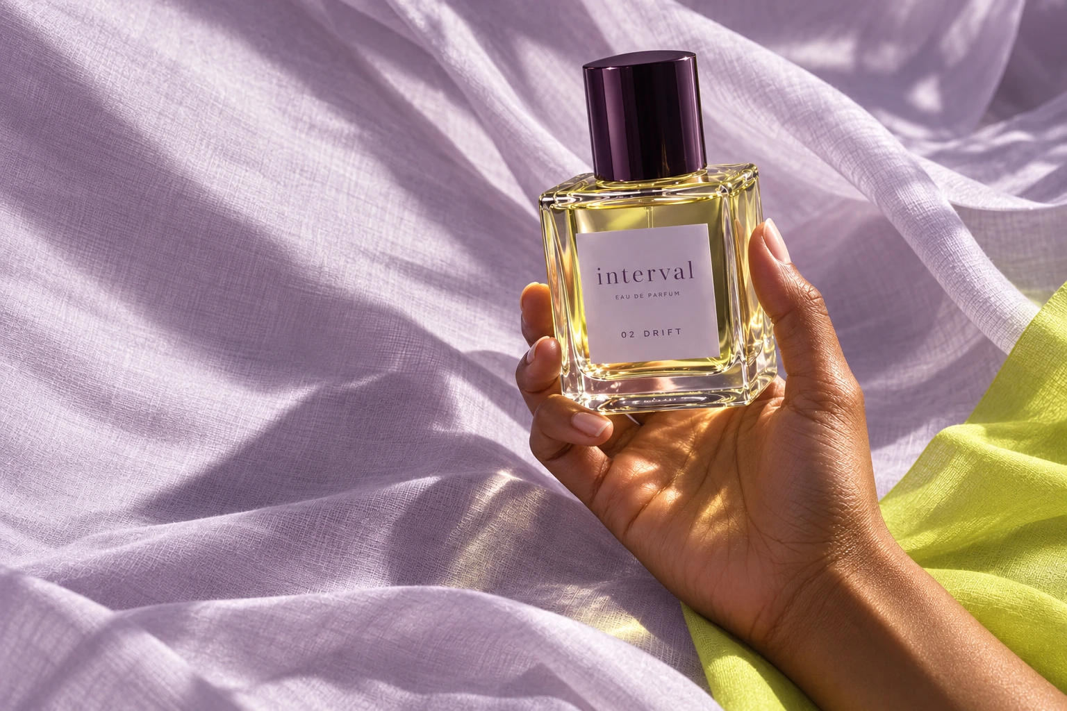 A hand holds INTERVAL Drift perfume against softly draped lilac fabric in natural sunlight